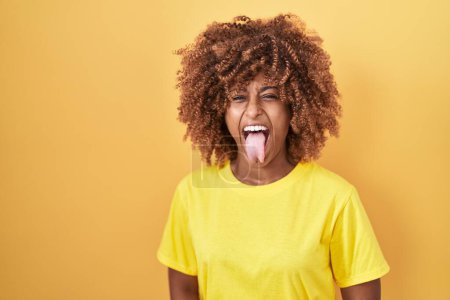 Photo for Young hispanic woman with curly hair standing over yellow background sticking tongue out happy with funny expression. emotion concept. - Royalty Free Image