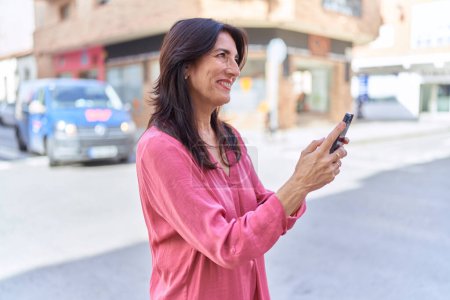 Foto de Mujer hispana de mediana edad sonriendo confiada usando smartphone en la calle - Imagen libre de derechos
