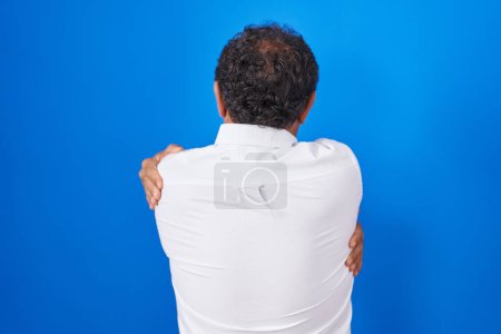 Foto de Hombre hispano de mediana edad de pie sobre fondo azul abrazándose feliz y positivo desde atrás. amor propio y cuidado personal - Imagen libre de derechos