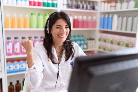 Téléchargez les photos : Middle age hispanic woman working at pharmacy drugstore using headset and computer screaming proud, celebrating victory and success very excited with raised arm - en image libre de droit
