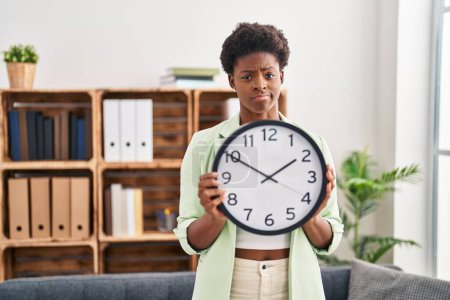 Foto de Mujer afroamericana sosteniendo gran reloj escéptico y nervioso, frunciendo el ceño molesto debido a un problema. persona negativa. - Imagen libre de derechos