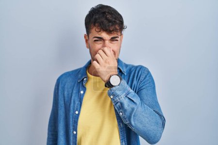 Téléchargez les photos : Jeune homme hispanique debout sur fond bleu sentant quelque chose de puant et dégoûtant, odeur intolérable, retenant souffle avec les doigts sur le nez. mauvaise odeur - en image libre de droit