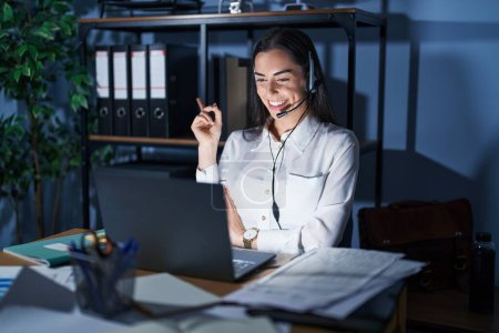 Photo for Young brunette woman wearing call center agent headset working late at night with a big smile on face, pointing with hand and finger to the side looking at the camera. - Royalty Free Image