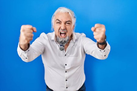 Photo for Middle age man with grey hair standing over blue background angry and mad raising fists frustrated and furious while shouting with anger. rage and aggressive concept. - Royalty Free Image