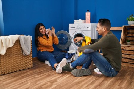 Foto de Hombre y mujer pareja sonriendo confiado jugando con la ropa en la lavandería - Imagen libre de derechos