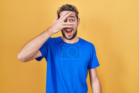 Foto de Hispanic man with beard standing over yellow background peeking in shock covering face and eyes with hand, looking through fingers with embarrassed expression. - Imagen libre de derechos