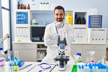 Foto de Young hispanic man with beard working at scientist laboratory looking confident at the camera with smile with crossed arms and hand raised on chin. thinking positive. - Imagen libre de derechos