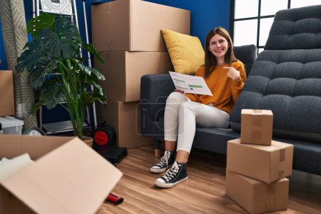 Photo for Young brunette woman sitting on the sofa at new home looking at documents pointing finger to one self smiling happy and proud - Royalty Free Image