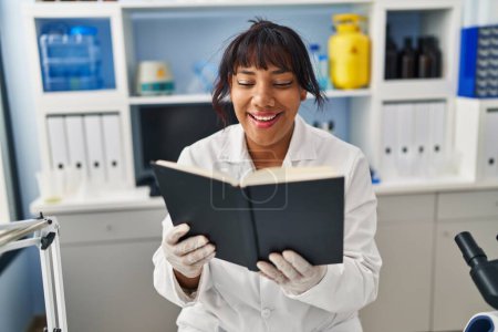 Foto de Mujer hispana trabajando en laboratorio científico leyendo un libro sonriendo y riendo en voz alta porque divertida broma loca. - Imagen libre de derechos