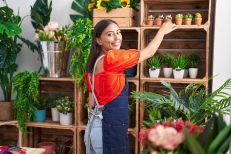 Foto de Joven hermosa mujer hispana florista sosteniendo planta de estanterías en floristería - Imagen libre de derechos