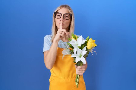 Foto de Young caucasian woman wearing florist apron holding flowers asking to be quiet with finger on lips. silence and secret concept. - Imagen libre de derechos