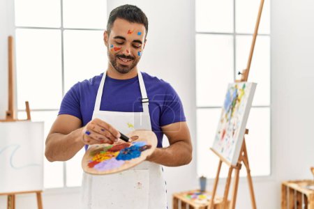 Téléchargez les photos : Jeune homme hispanique souriant confiant mélanger la couleur sur la palette au studio d'art - en image libre de droit