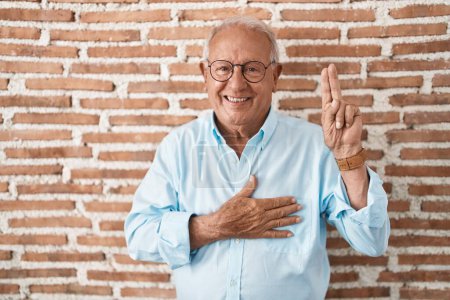 Photo for Senior man with grey hair standing over bricks wall smiling swearing with hand on chest and fingers up, making a loyalty promise oath - Royalty Free Image