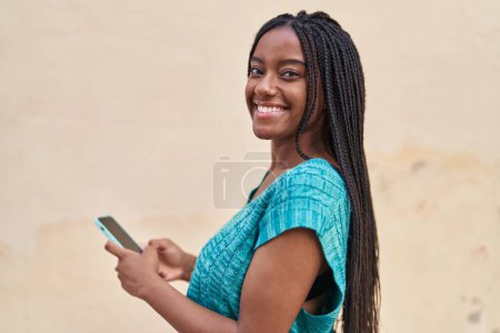 Téléchargez les photos : Afro-américaine femme souriant confiant en utilisant smartphone à la rue - en image libre de droit