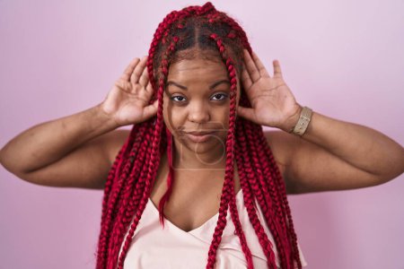 Foto de Mujer afroamericana con el pelo trenzado de pie sobre fondo rosa tratando de escuchar ambas manos en el gesto de la oreja, curioso por chismes. problema auditivo, sordo - Imagen libre de derechos