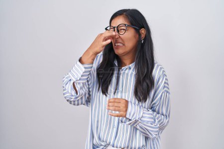 Téléchargez les photos : Jeune femme hispanique portant des lunettes sentant quelque chose de puant et dégoûtant, odeur intolérable, retenant son souffle avec les doigts sur le nez. mauvaise odeur - en image libre de droit