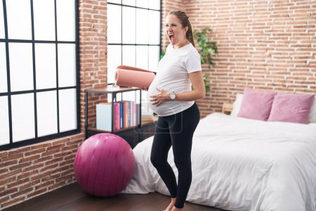 Téléchargez les photos : Young pregnant woman holding pilate mat at bedroom angry and mad screaming frustrated and furious, shouting with anger. rage and aggressive concept. - en image libre de droit