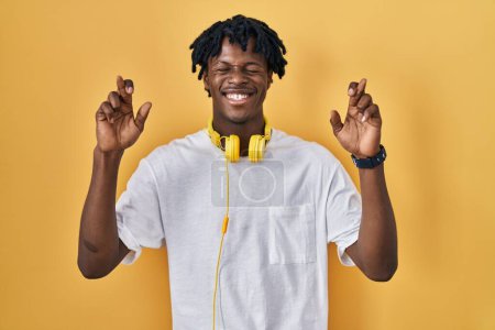 Foto de Joven hombre africano con rastas de pie sobre fondo amarillo gesto dedo cruzado sonriendo con esperanza y los ojos cerrados. suerte y concepto supersticioso. - Imagen libre de derechos