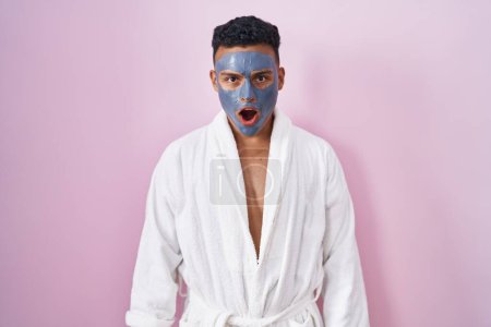 Téléchargez les photos : Young hispanic man wearing beauty face mask and bath robe in shock face, looking skeptical and sarcastic, surprised with open mouth - en image libre de droit