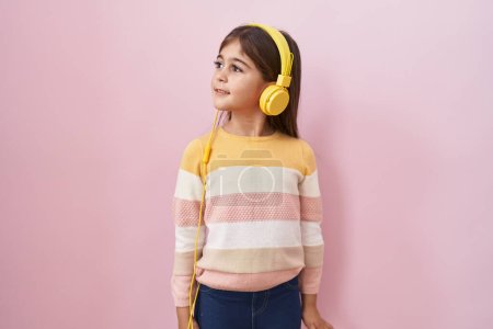 Téléchargez les photos : Petite fille hispanique écoutant de la musique à l'aide d'écouteurs regardant vers le côté, pose de profil relax avec le visage naturel et sourire confiant. - en image libre de droit