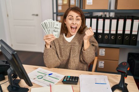 Foto de Young beautiful woman working at small business ecommerce holding money screaming proud, celebrating victory and success very excited with raised arms - Imagen libre de derechos