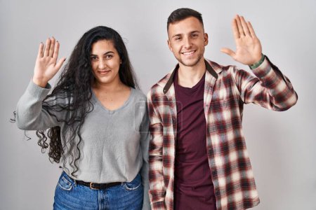 Photo for Young hispanic couple standing over white background waiving saying hello happy and smiling, friendly welcome gesture - Royalty Free Image