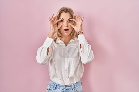 Téléchargez les photos : Jeune femme caucasienne portant une chemise blanche décontractée sur fond rose essayant d'ouvrir les yeux avec les doigts, endormie et fatiguée pour la fatigue matinale - en image libre de droit