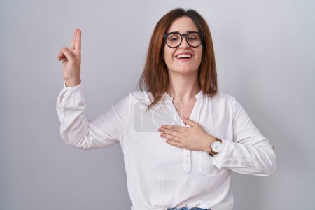 Foto de Brunette woman standing over white isolated background smiling swearing with hand on chest and fingers up, making a loyalty promise oath - Imagen libre de derechos
