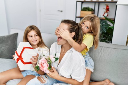 Foto de Madre e hijas sonriendo sorpresa segura con regalo y flores en casa - Imagen libre de derechos