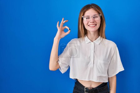 Photo for Beautiful woman standing over blue background smiling positive doing ok sign with hand and fingers. successful expression. - Royalty Free Image