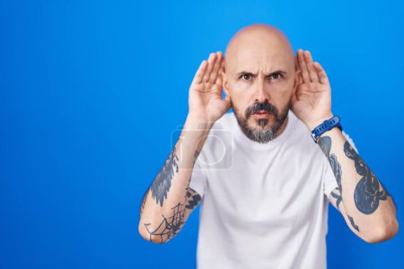 Téléchargez les photos : Homme hispanique avec des tatouages debout sur fond bleu essayant d'entendre les deux mains sur le geste de l'oreille, curieux de commérages. problème auditif, sourd - en image libre de droit
