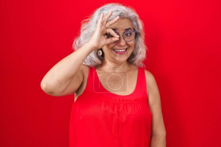Foto de Middle age woman with grey hair standing over red background doing ok gesture with hand smiling, eye looking through fingers with happy face. - Imagen libre de derechos