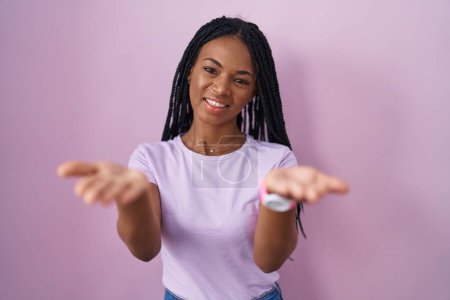 Téléchargez les photos : Femme afro-américaine avec des tresses debout sur fond rose souriant joyeuse offrant des mains donnant assistance et acceptation. - en image libre de droit