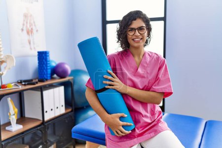 Téléchargez les photos : Jeune femme latine portant un uniforme de physiothérapeute tenant un tapis de yoga à la clinique - en image libre de droit