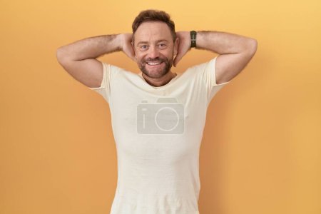 Téléchargez les photos : Homme d'âge moyen avec barbe debout sur fond jaune relaxant et étirant, bras et mains derrière la tête et le cou souriant heureux - en image libre de droit