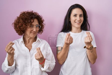 Téléchargez les photos : Hispanic mother and daughter wearing casual white t shirt over pink background doing money gesture with hands, asking for salary payment, millionaire business - en image libre de droit