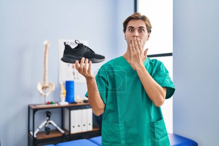 Photo for Young man working at physiotherapy clinic holding shoe covering mouth with hand, shocked and afraid for mistake. surprised expression - Royalty Free Image