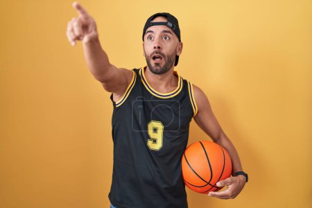 Photo for Middle age bald man holding basketball ball over yellow background pointing with finger surprised ahead, open mouth amazed expression, something on the front - Royalty Free Image