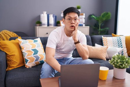 Foto de Young asian man using laptop at home sitting on the sofa afraid and shocked, surprise and amazed expression with hands on face - Imagen libre de derechos