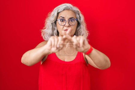 Foto de Mujer de mediana edad con el pelo gris de pie sobre el fondo rojo expresión rechazo cruzando los dedos haciendo signo negativo - Imagen libre de derechos