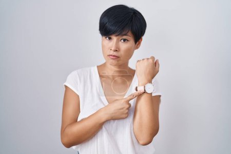 Photo for Young asian woman with short hair standing over isolated background in hurry pointing to watch time, impatience, looking at the camera with relaxed expression - Royalty Free Image