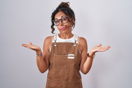 Photo for Middle age woman wearing apron over white background clueless and confused expression with arms and hands raised. doubt concept. - Royalty Free Image