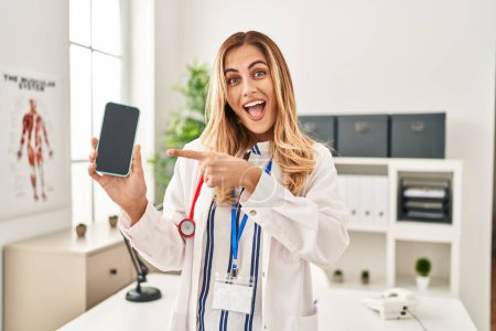 Téléchargez les photos : Jeune femme blonde médecin travaillant à la clinique montrant écran smartphone souriant heureux pointant avec la main et le doigt - en image libre de droit