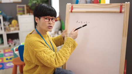 Photo for Young chinese woman preschool teacher sitting on chair writing on chalkboard at kindergarten - Royalty Free Image