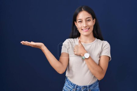Foto de Mujer hispana joven de pie sobre fondo azul mostrando la palma de la mano y haciendo buen gesto con los pulgares hacia arriba, sonriendo feliz y alegre - Imagen libre de derechos