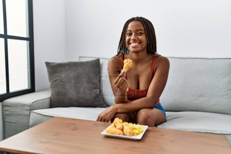 Foto de Joven afroamericana americana comiendo pollo frito sentada en sofá en casa - Imagen libre de derechos