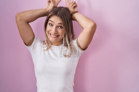 Foto de Blonde caucasian woman standing over pink background posing funny and crazy with fingers on head as bunny ears, smiling cheerful - Imagen libre de derechos