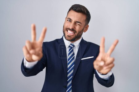 Foto de Hombre hispano guapo vistiendo traje y corbata sonriendo con la lengua hacia fuera mostrando los dedos de ambas manos haciendo signo de victoria. número dos. - Imagen libre de derechos