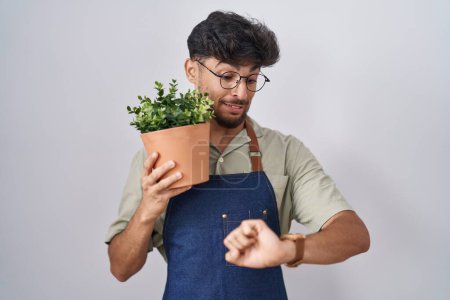 Téléchargez les photos : Homme arabe avec barbe tenant pot de plante verte regardant l'heure de la montre inquiet, peur de se faire tard - en image libre de droit