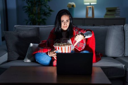 Photo for Hispanic woman eating popcorn watching a movie on the sofa looking unhappy and angry showing rejection and negative with thumbs down gesture. bad expression. - Royalty Free Image